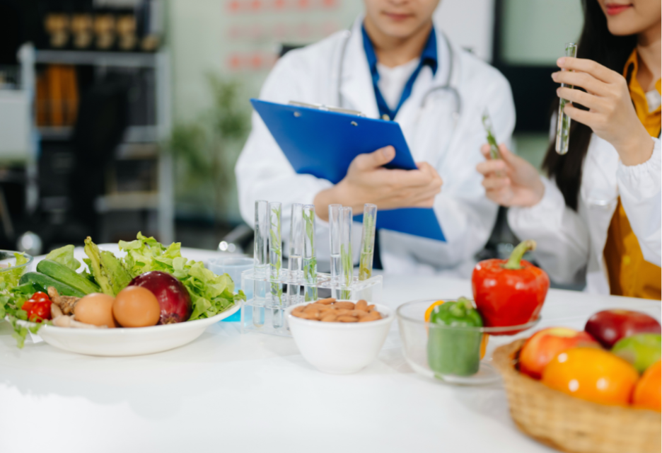 A laboratory scene showing a healthcare professional reviewing notes while another person examines test tubes, with fresh fruits and vegetables displayed on the table in the foreground.